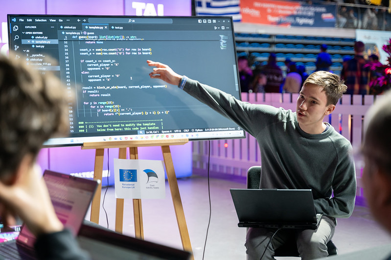 Õpilane näitab programmeerimiskoodi suurel ekraanil, esiplaanil osalejad ja taustal nähtav Euroopa Liidu logo ning Robotexi võistluskeskkond.