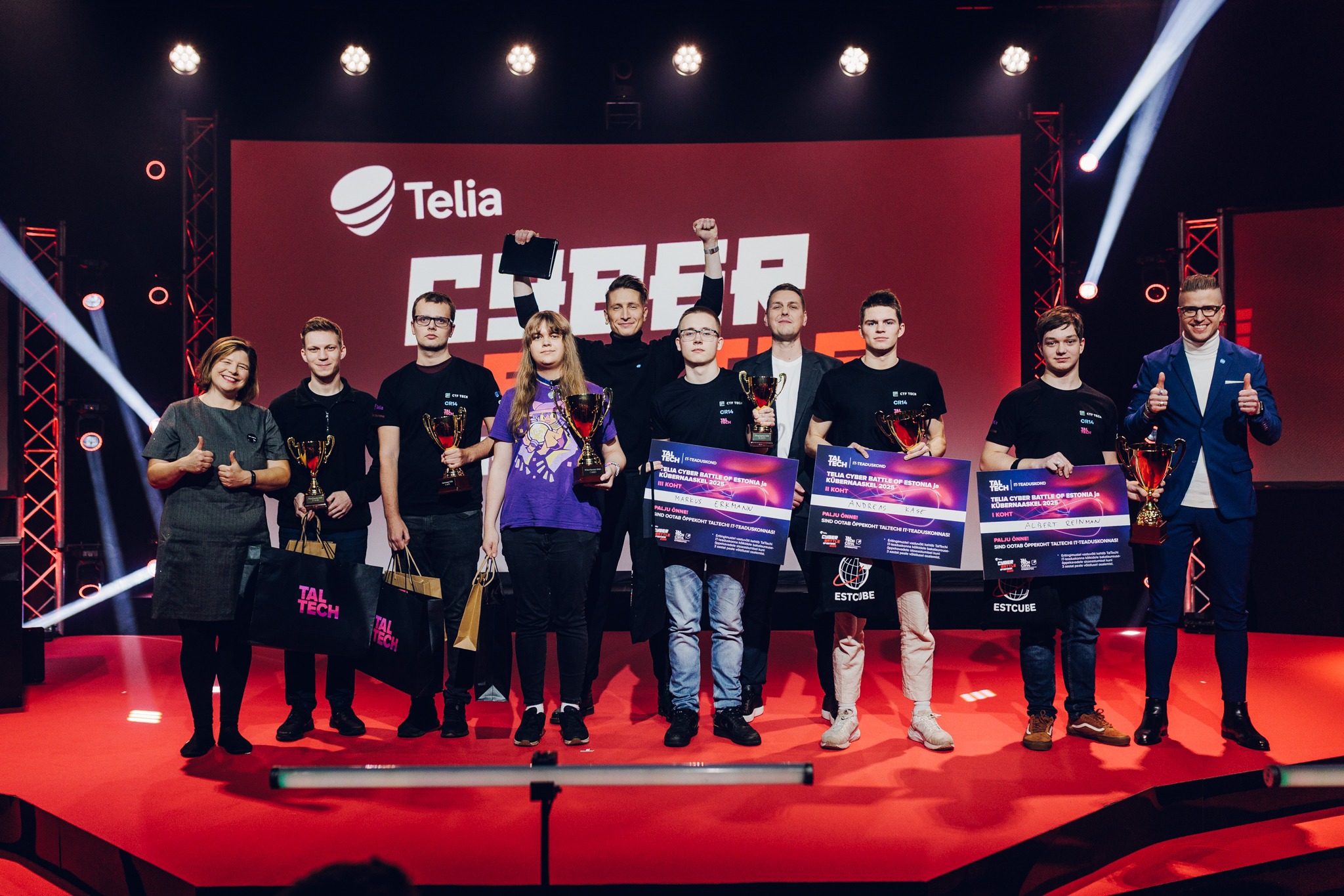 Telia Cyber Battle ja CyberSpike 2025 võistluse võitjad ja auhinnasaajad seisavad punase lava peal trofeede ja auhinnatšekkidega. Taustaks on suur Telia logoga ekraan ning lava on valgustatud professionaalsete valgustega. Võistkonnad kannavad TalTechi ja võistluse särke.