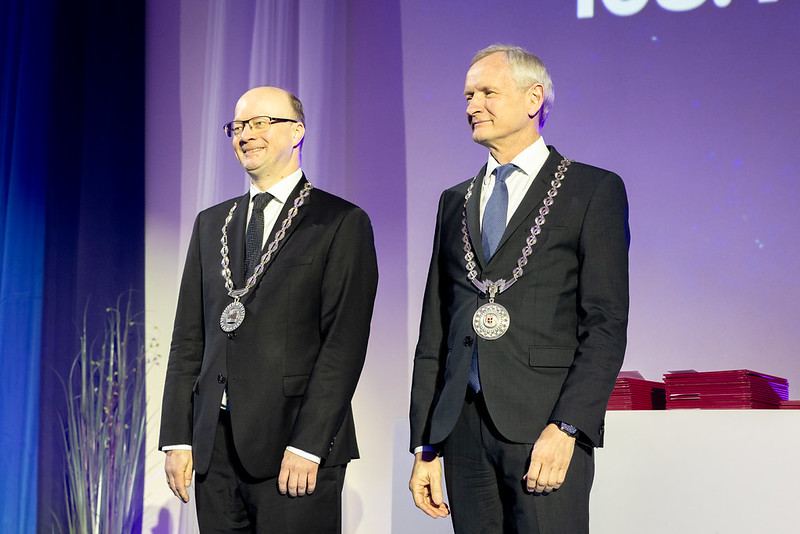 Kaks meest seisavad laval pidulikus keskkonnas, mõlemad kannavad tumedaid ülikondi ja kaelas suuri hõbedasi ametikette medaliga. Nad naeratavad kergelt ja vaatavad publiku suunas. Taustal on ekraan ning kõrval laual on virn punaseid kaustu.