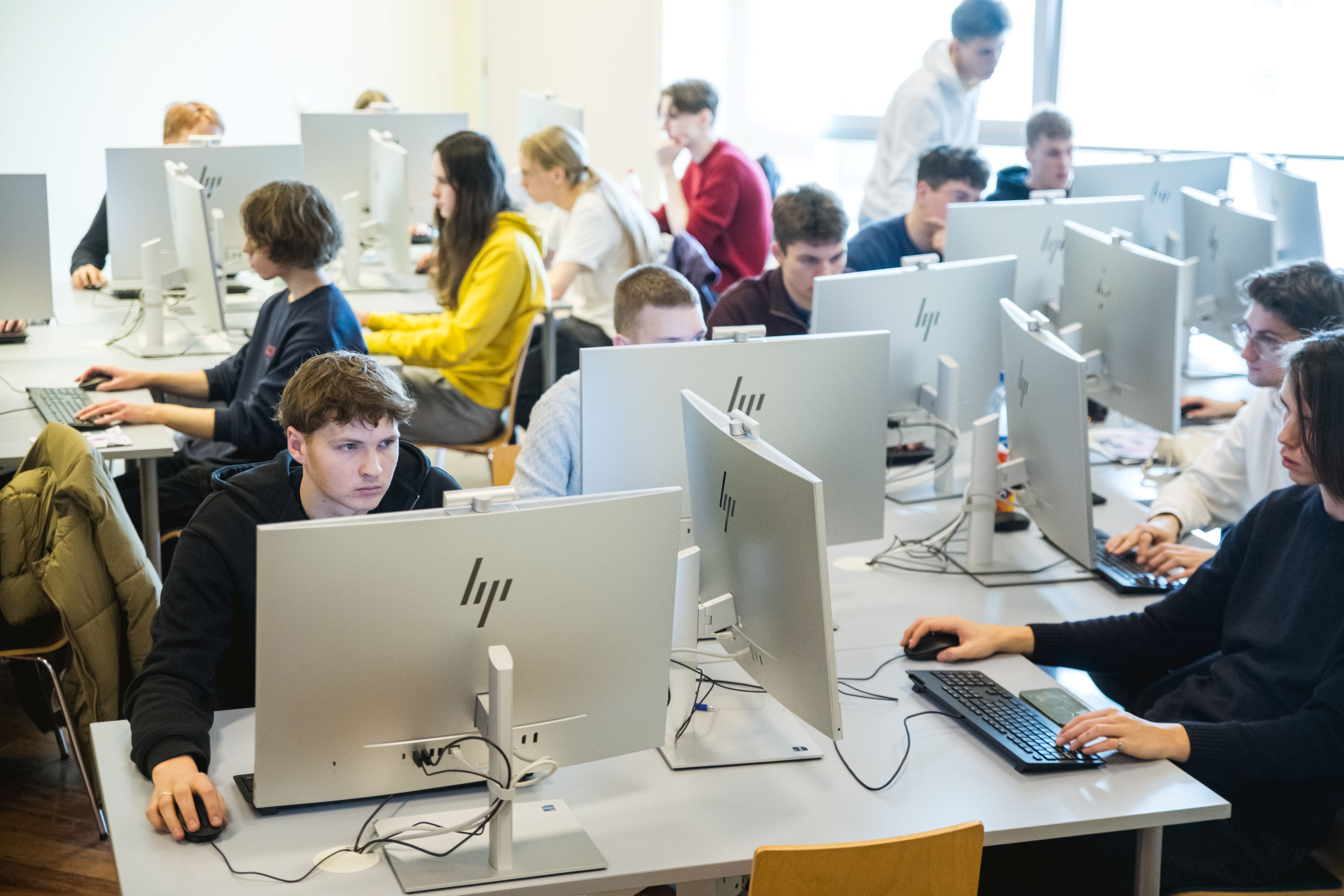 Arvutiklassis töötab rühm tudengeid lauaarvutites. Noored istuvad monitoride taga ja keskenduvad programmeerimisele või muudele IT-ülesannetele.