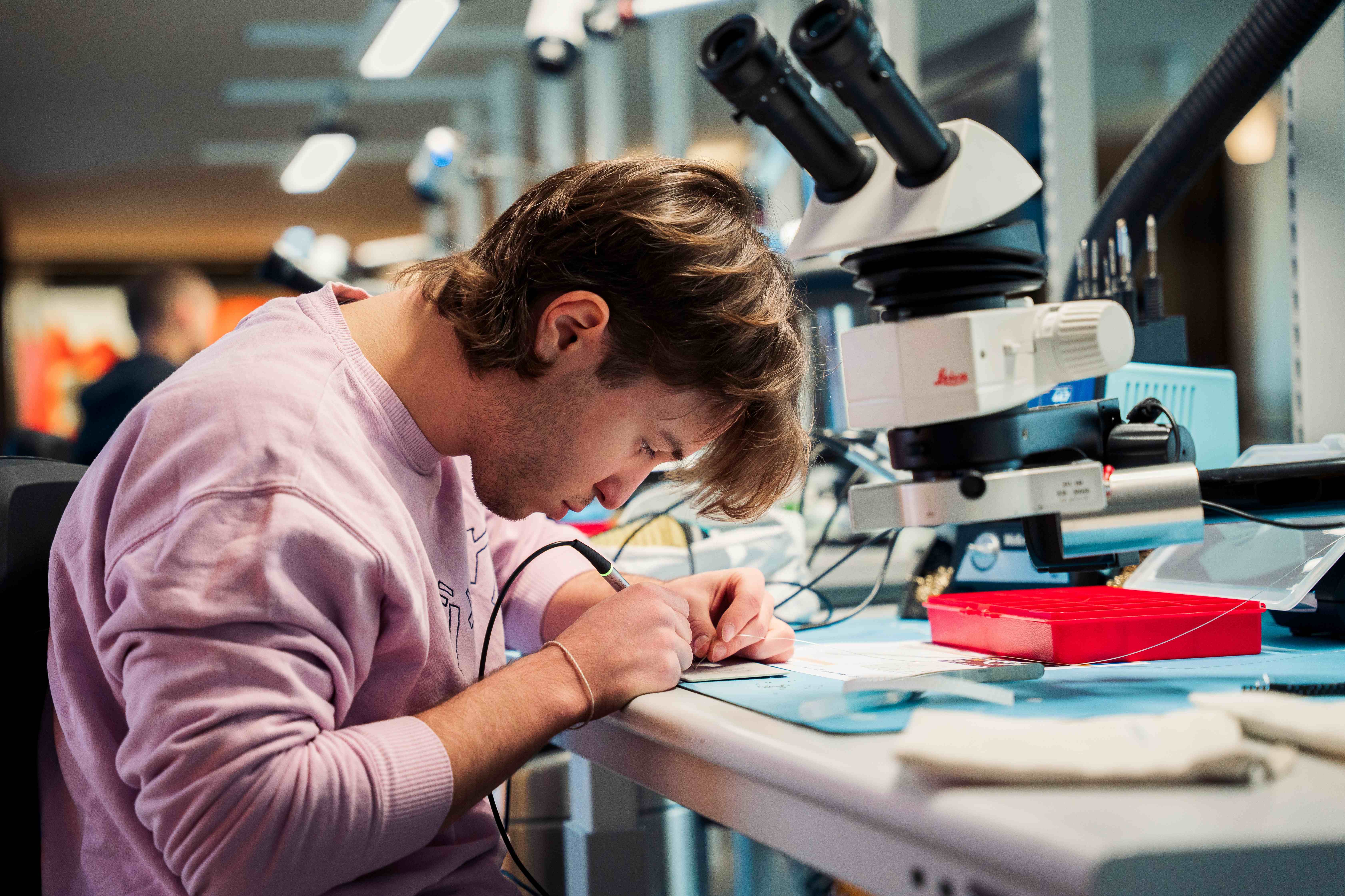 Tudeng töötab mikroelektroonika laboris täppisseadme kallal. Töölaual on mikroskoop ja elektroonikaseadmed, mille kõrval ta keskendunult jootmis- või mõõtetööd teeb.