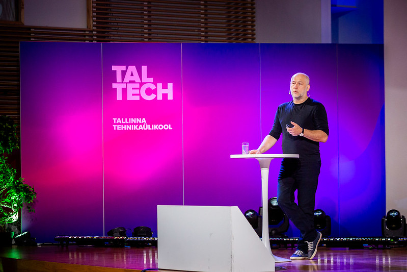 Mees esineb laval TalTechi taustaga, seistes väikese kõrglaua kõrval ja rääkides publikule; taustal on värviline valguslahendus ja tekst „TalTech Tallinna Tehnikaülikool“.