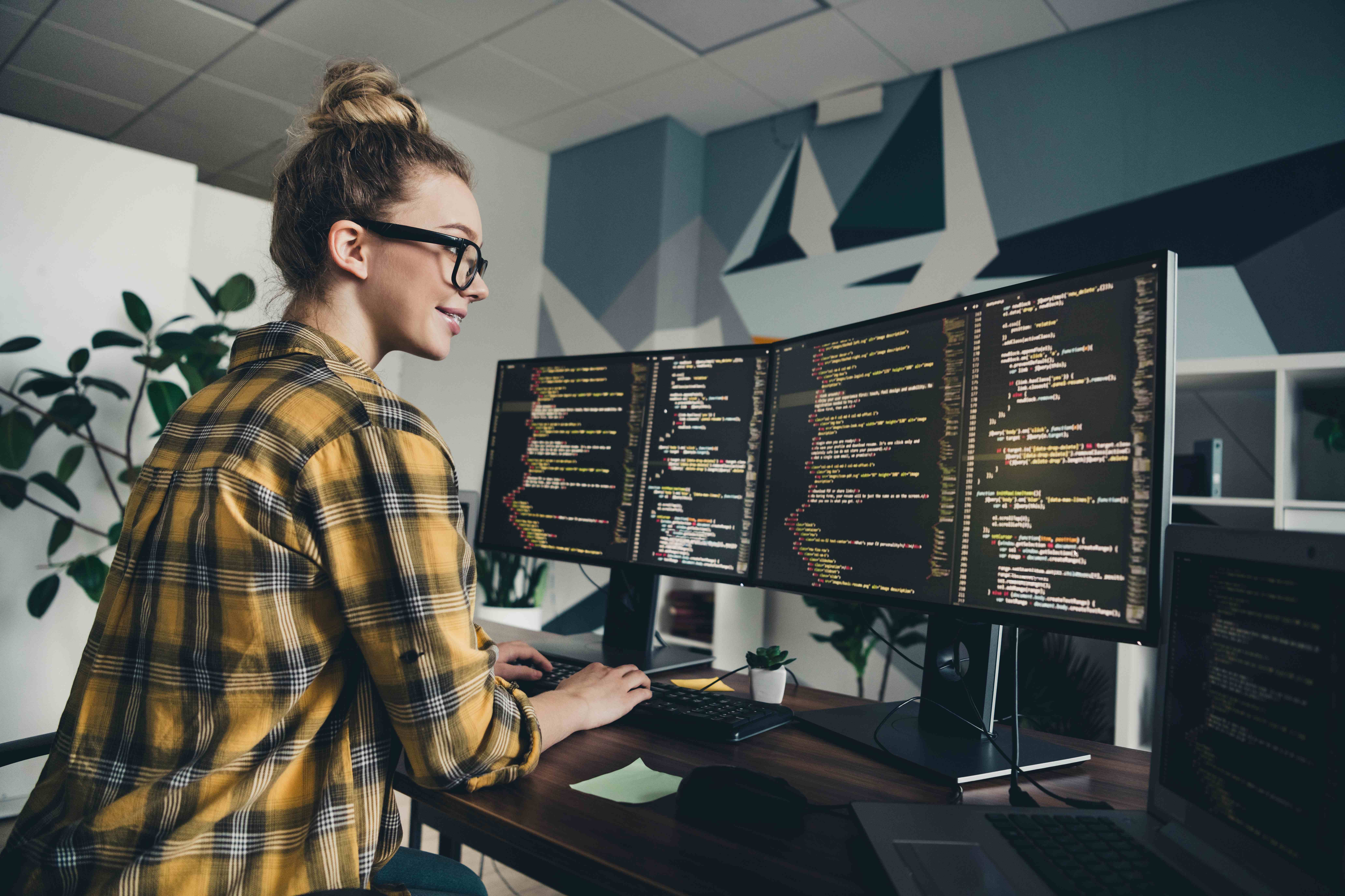 Noor naine prillides ja kollases ruudukleidis töötab kaasaegses kontoris kahe suure monitori taga, mille ekraanidel on näha programmeerimiskoodi. Taustal on taimi ja modernne seinakujundus.