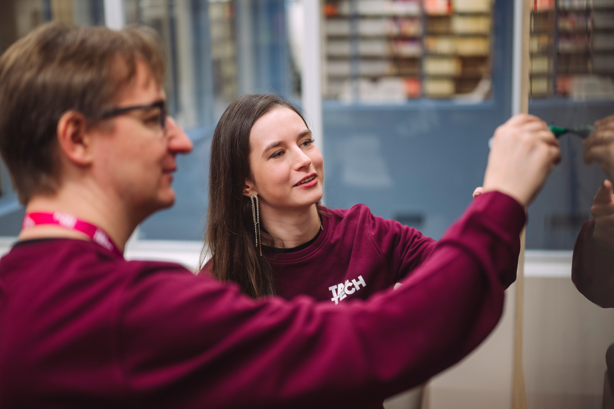 Naine ja mees TalTechi bordoo värvi üliõpilaspusades kirjutavad auditooriumis tahvlile, naine naeratab ja vaatab kõrvale mehe poole.