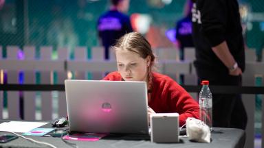 Noor naissoost võistleja punases kampsus töötab HP sülearvutiga võistluslaual, taustal näha Robotexi värvilisi tulesid ja teisi osalejaid