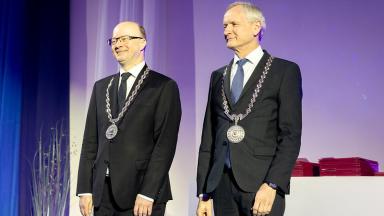 Kaks meest seisavad laval pidulikus keskkonnas, mõlemad kannavad tumedaid ülikondi ja kaelas suuri hõbedasi ametikette medaliga. Nad naeratavad kergelt ja vaatavad publiku suunas. Taustal on ekraan ning kõrval laual on virn punaseid kaustu.