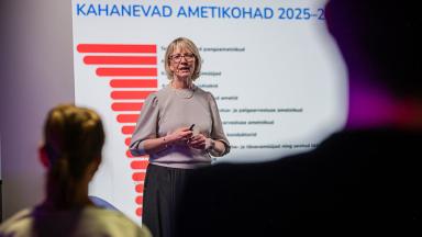 Esineja seisab laval ja peab ettekannet tuleviku tööturu teemal. Taustal oleval slaidil on pealkiri „Kahanevad ametikohad 2025–…“ ning punane tulpdiagramm. Esiplaanil on näha kuulajate siluetid.