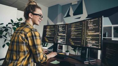 Noor naine prillides ja kollases ruudukleidis töötab kaasaegses kontoris kahe suure monitori taga, mille ekraanidel on näha programmeerimiskoodi. Taustal on taimi ja modernne seinakujundus.