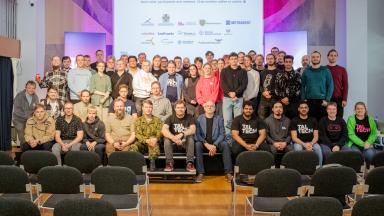 Fly or Die drooni­häkatoni avapäeva grupifoto TalTechis – osalejad, mentorid ja korraldajad koos laval, taustal ekraan ürituse logodega.