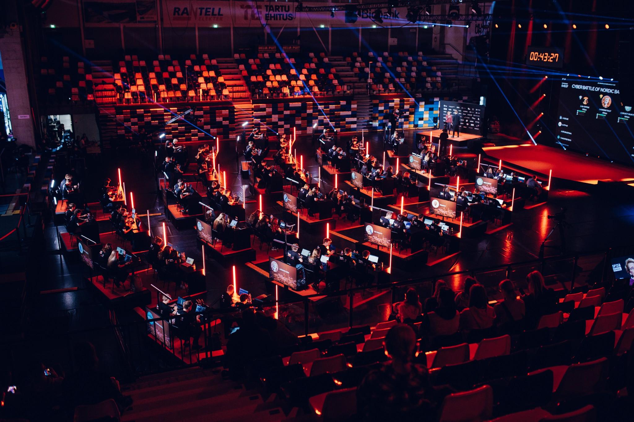 Suur küberturvalisuse võistlus saalis — arvutilauad valguspulkade vahel, kümned osalejad ekraanide taga, taustal laval suur ekraan.