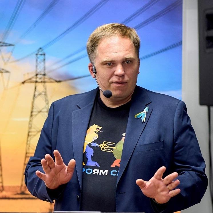 Mees esinemas, peas mikrofoniga peakomplekt ja seljas tume pintsak ning trükiga T-särk. Ta teeb kätega selgitavat žesti. Taustal on elektriliinide kujutis, viidates tehnoloogia või energia teemalisele ettekandele.
