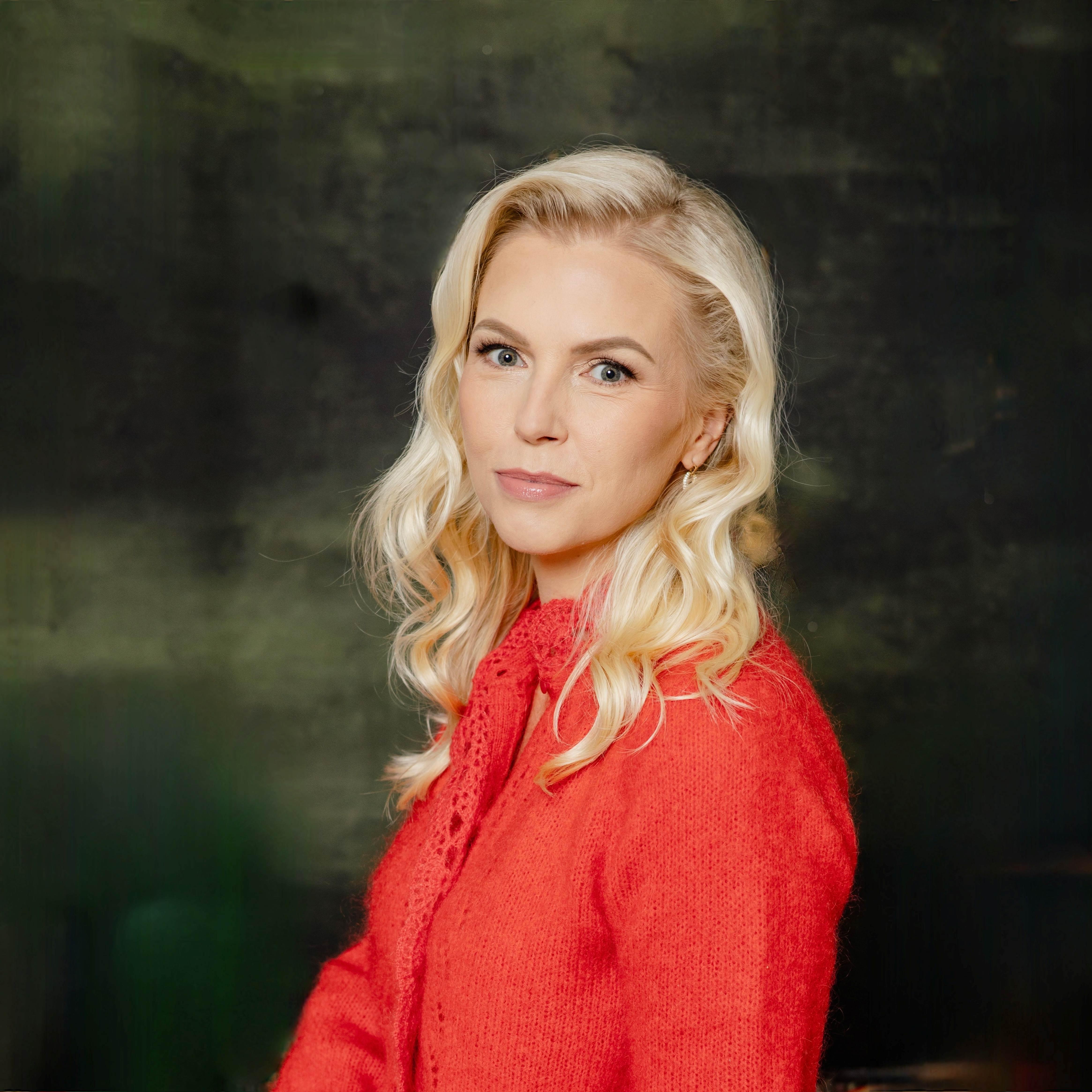 Portreefoto naisest, kes vaatab kaamerasse. Tal on pikad heledad lainelised juuksed ja seljas erkpunane kampsun. Taust on tumedates toonides ja pehmelt hägustatud, rõhutades portree isikupära.
