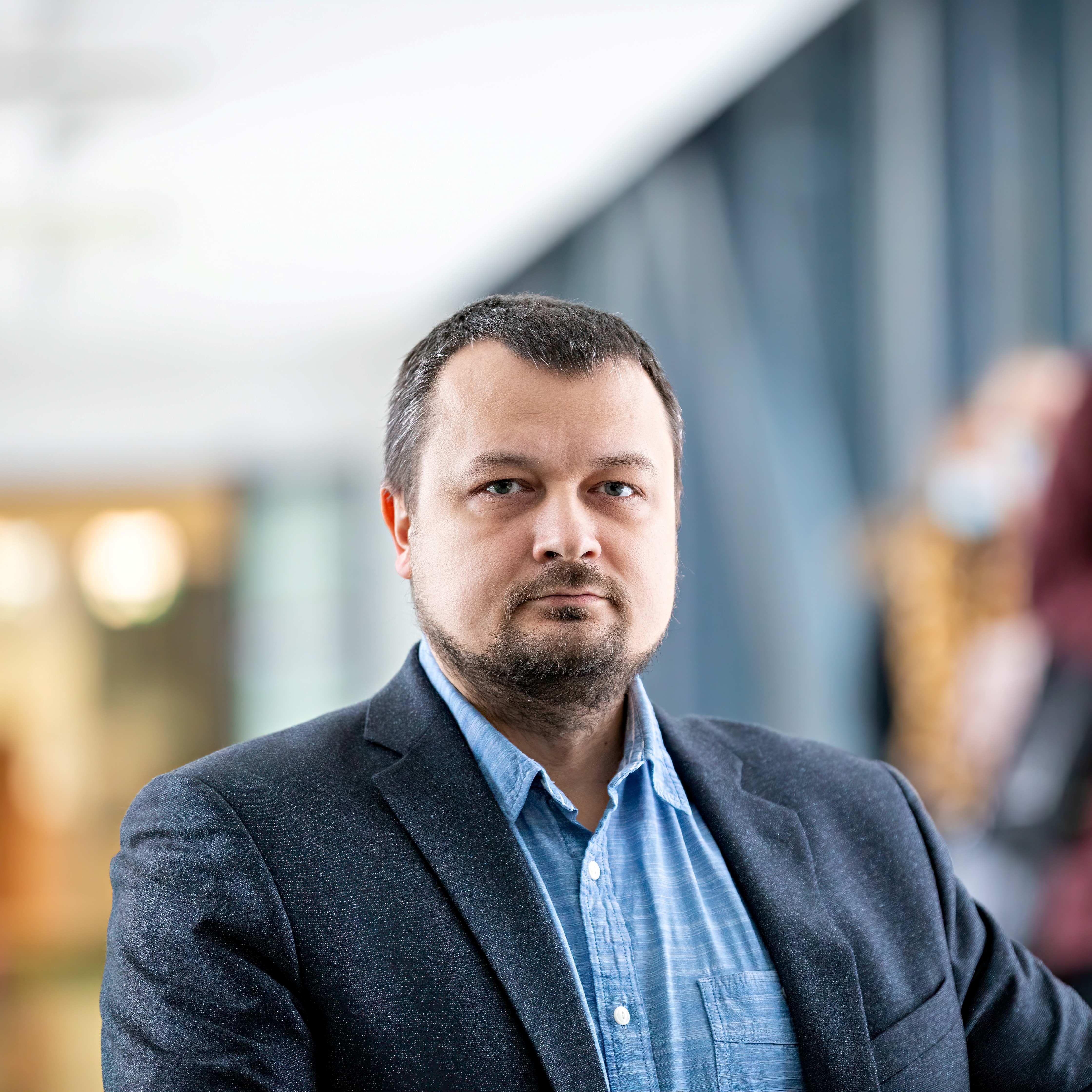 Lühikese soengu ja pöetud habemega mees vaatab tõsise ilmega kaamerasse. Taustal on hele lagi ja tumehall sein.
