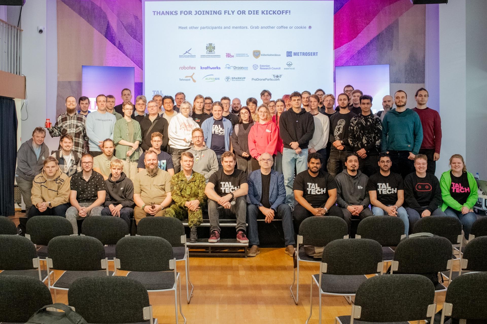 Group photo from the opening day of the Fly or Die drone hackathon at TalTech — participants, mentors and organisers on stage with the event logos displayed on screen behind them.