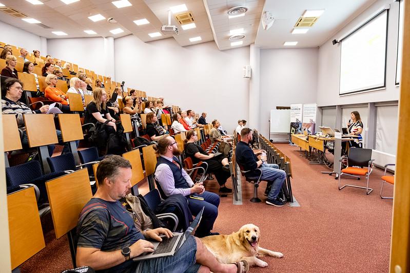 IT-õpetajate suvekooli loeng TalTechi IT Kolledži auditooriumis; publik kuulab esinejat, esiplaanil mees sülearvutiga ja tema kõrval lamab koer.