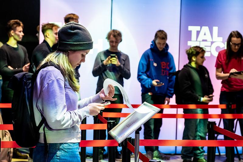 TalTechi AI-võistluse 2025 osaleja uurib paberit ülesande lahendamiseks, taustal teised osalejad telefonidega